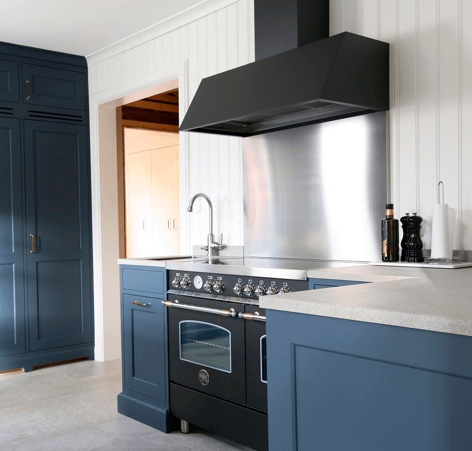 Vägghängda köksfläkten Aero i svart utförande skapar kontrast mot stänkskydd i rostfritt och ljust kakel i modernt kök med blåa köksluckor.