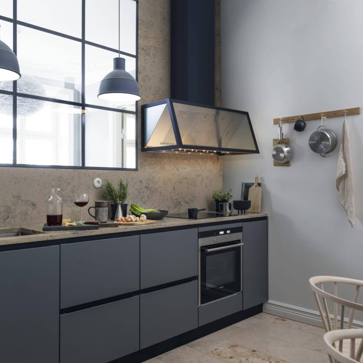 Vägghängda köksfläkten Fasett i svart utförande i modern köksmiljö med industrifönster och gråblåa köksluckor.